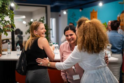 A group of women engage in friendly conversation at a lively event, surrounded by greenery and colorful decor