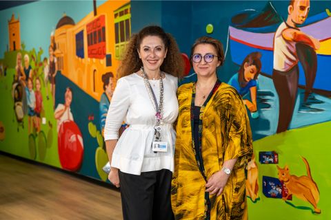 Two women pose together in front of a vibrant mural depicting cultural scenes and playful characters