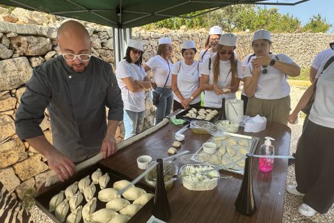 A chef demonstrates dumpling preparation while a group of people in white attire observes, surrounded by a rustic stone wall.