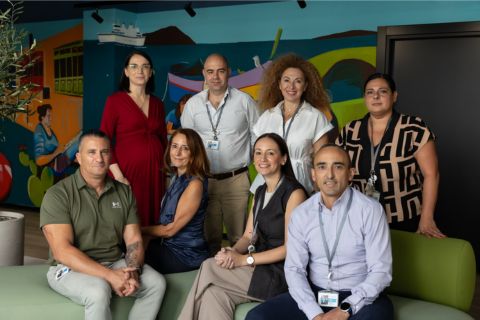 A diverse group of seven individuals poses together on a green couch in front of a vibrant mural