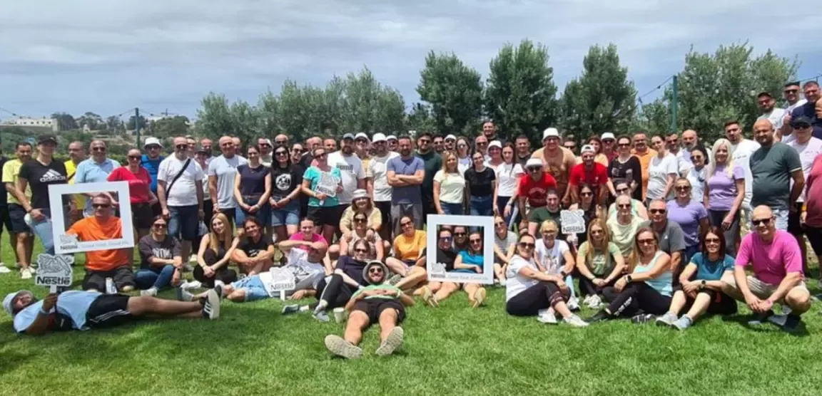 Large group of people posing outdoors on grass, some holding Nestlé signs and photo frames, with trees and a cloudy sky in the background.