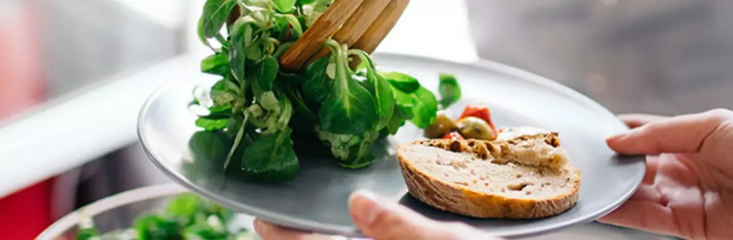 A hand holds a gray plate with fresh lettuce, olives, cherry tomatoes, and a slice of crusty bread
