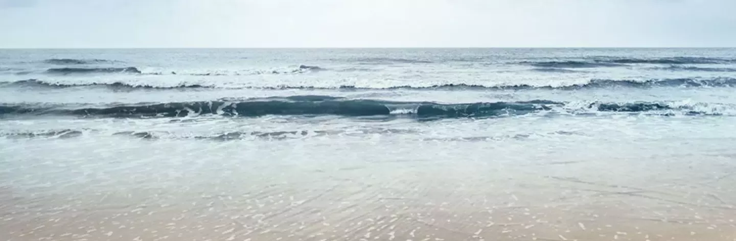 A tranquil seaside view featuring gentle waves lapping against a light sandy shore under a cloudy sky.