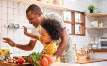 Father and child cooking together in a bright kitchen, reaching toward vegetables and tomatoes on the counter.