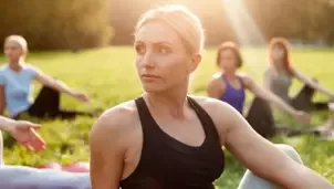 Blonde woman in black athletic wear sits outdoors in sunlight, with a blurred group practicing yoga on a grassy field behind her.