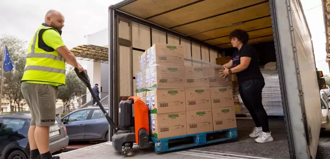 Workers unloading boxes of Nestlé products from a delivery truck at a loading dock, with trees and vehicles in the background.