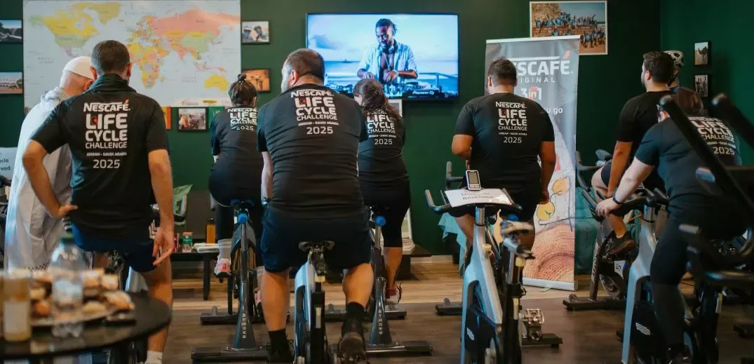 A group of participants on stationary bikes engaged in the Nescafé Life Cycle Challenge 2025, with a TV playing in the background.