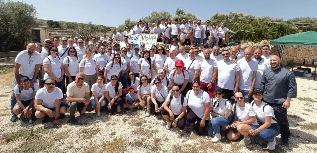 A large group of people in white shirts gathers outdoors, holding a sign for the Maya Foundation, under a clear blue sky