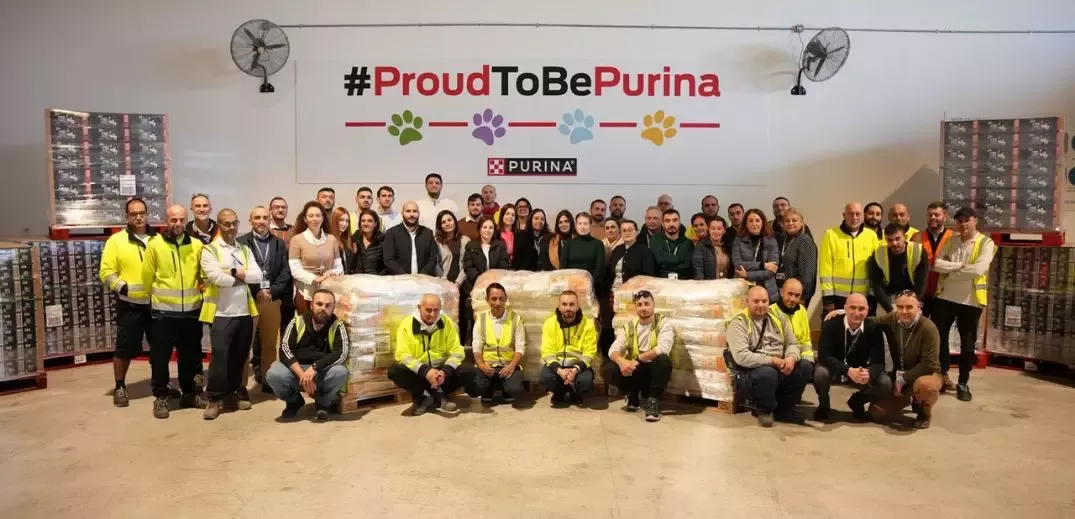 A group photo of Purina employees in a warehouse, standing in front of a "#ProudToBePurina" sign, with pallets of products in the background